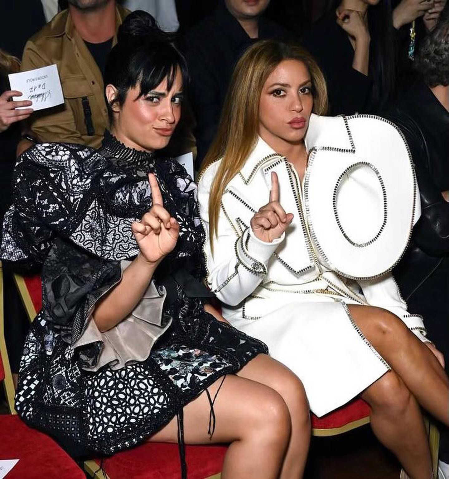 Dos mujeres sentadas haciendo un gesto de "no" con un dedo, mabas vestidas con trajes de alta costura, la primera lleva un traje oscuro con brillos y la segunda, una chaqueta larga banca con una inervención que dice NO.
