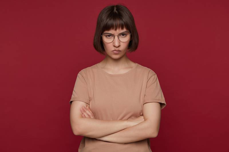 Mujer con pelo corto y lentes, de polera rosada, cruzando sus brazos y haciendo una cara de enojada.
