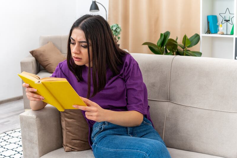 Imagen de mujer sentada en su sillón mientras lee un libro con cara de preocupada.