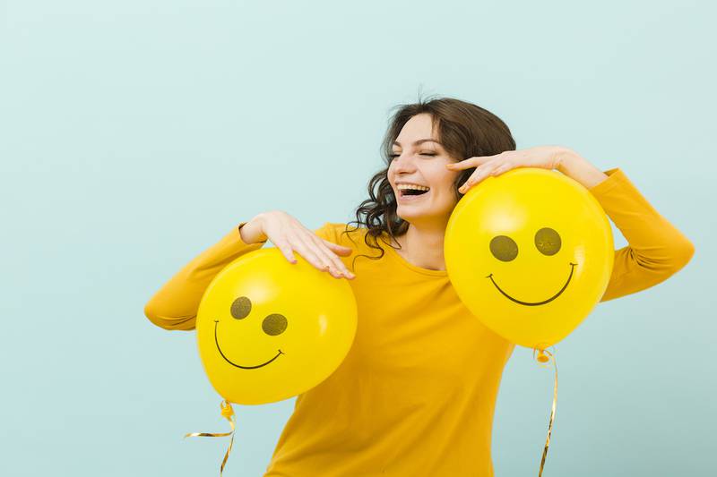 mujer de amarillo riéndose, con dos globos del mismo color que tienen dibujados unas sonrisas.