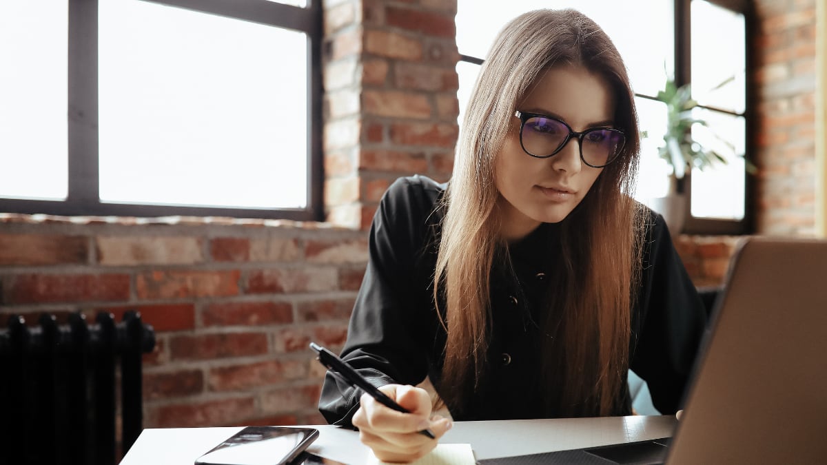 Mujer en una oficina, mirando el computador y anotando algo en un cuaderno.