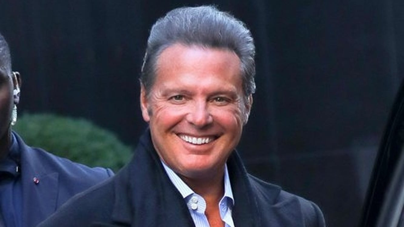 Hombre de avanzada edad sonriendo a la cámara con un abrigo y una bufanda negra y abajo tiene una camisa elegante abierta arriba