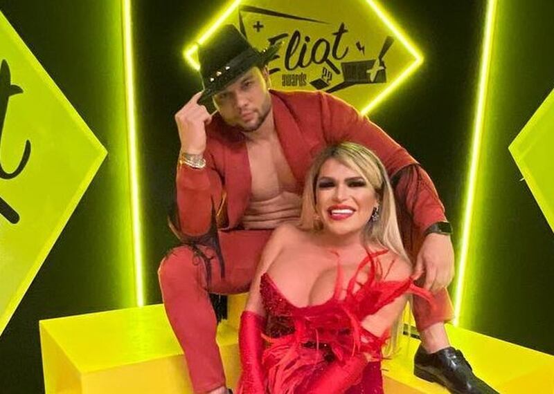Ambos sentados, webdy usa un vestido rojo sin tirante y Marlon un sombrero negro con un traje azul y sin camisa que deja ver su pecho musculado